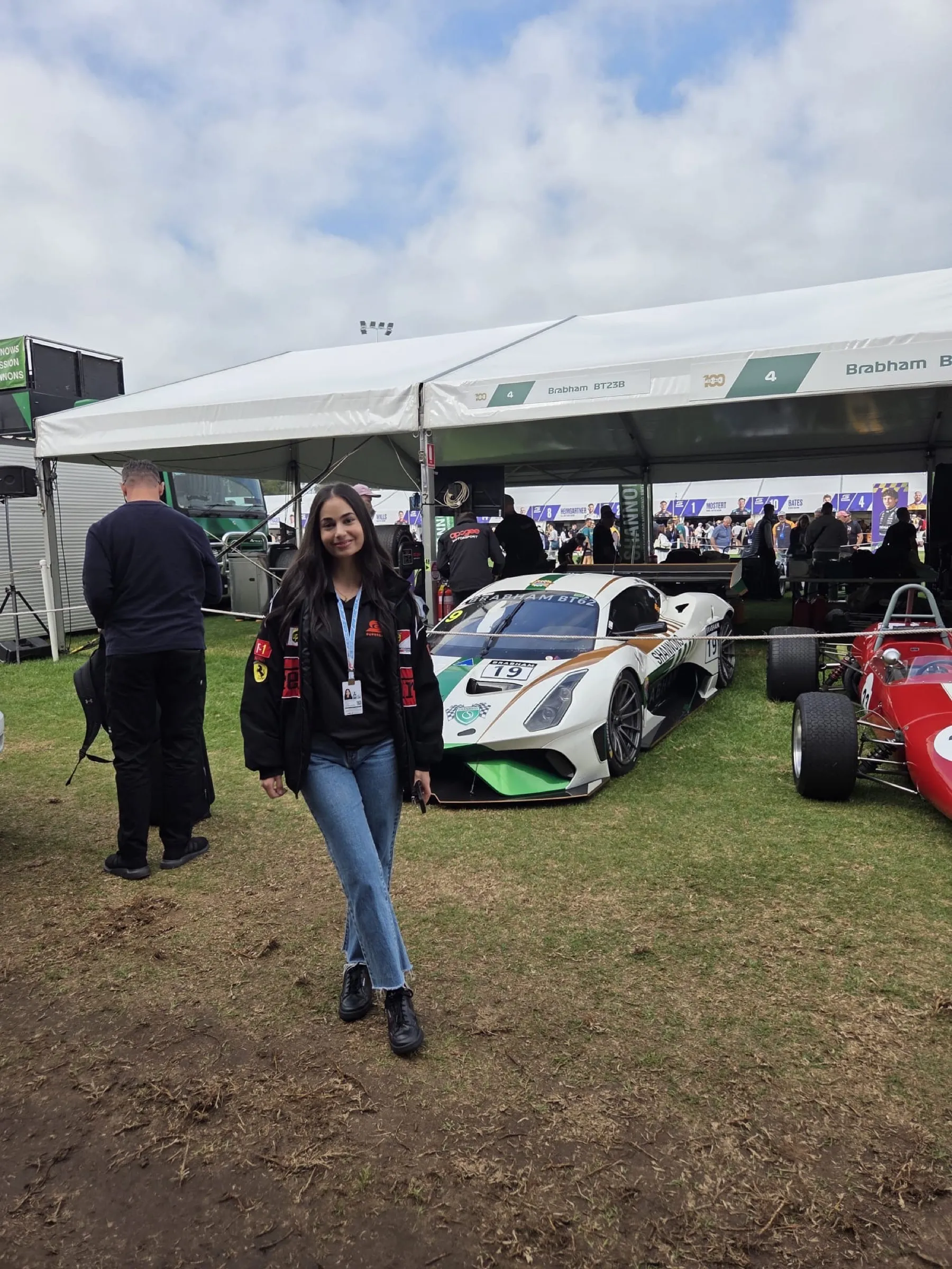 Maya standing on the grass at the Grand Prix wearing a lanyard and a black jacket, with a white racing car under a tent and other race cars behind her.