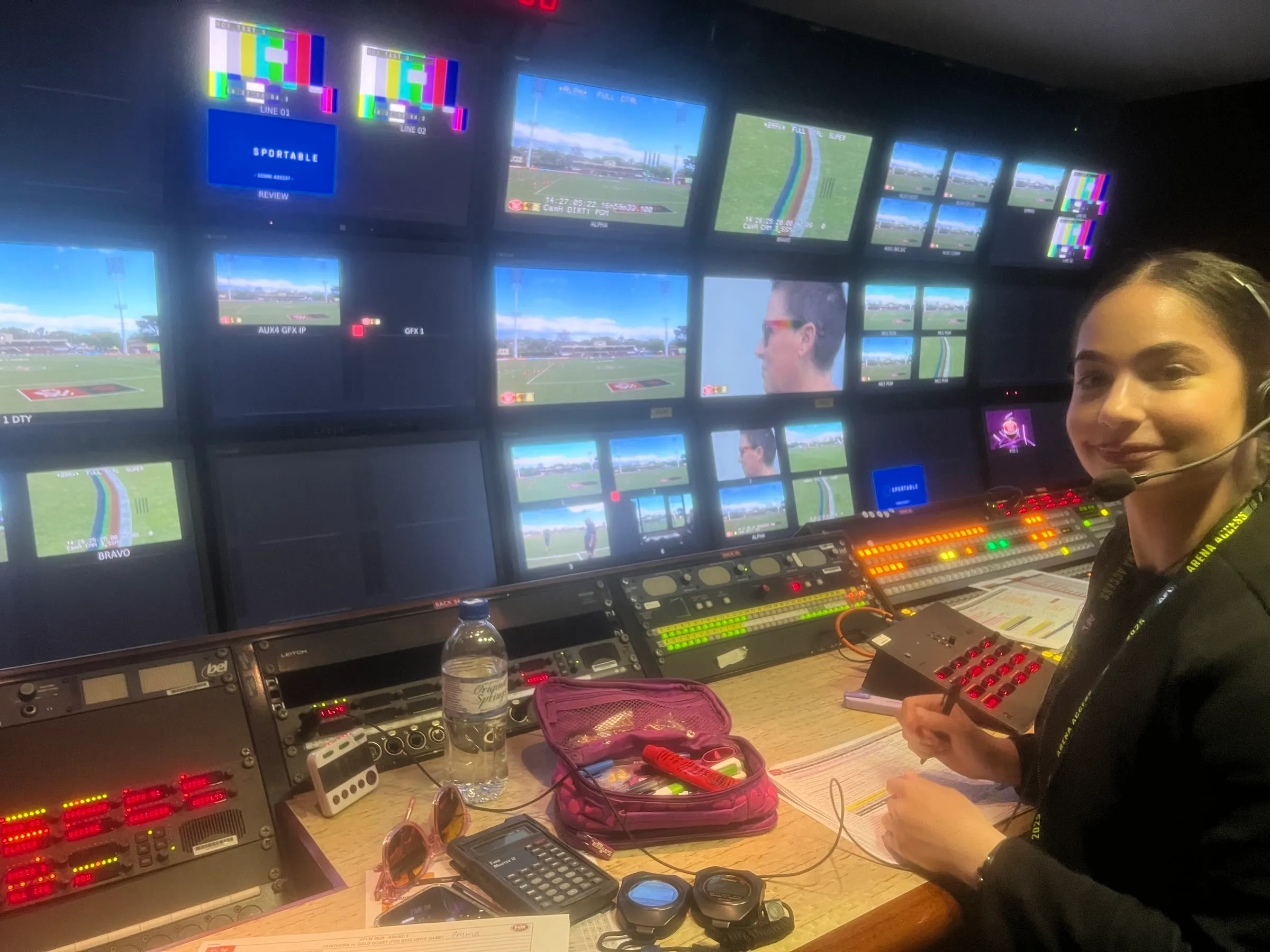 Maya wearing a headset in front of a wall of live sport monitors in an outside-broadcast control room.