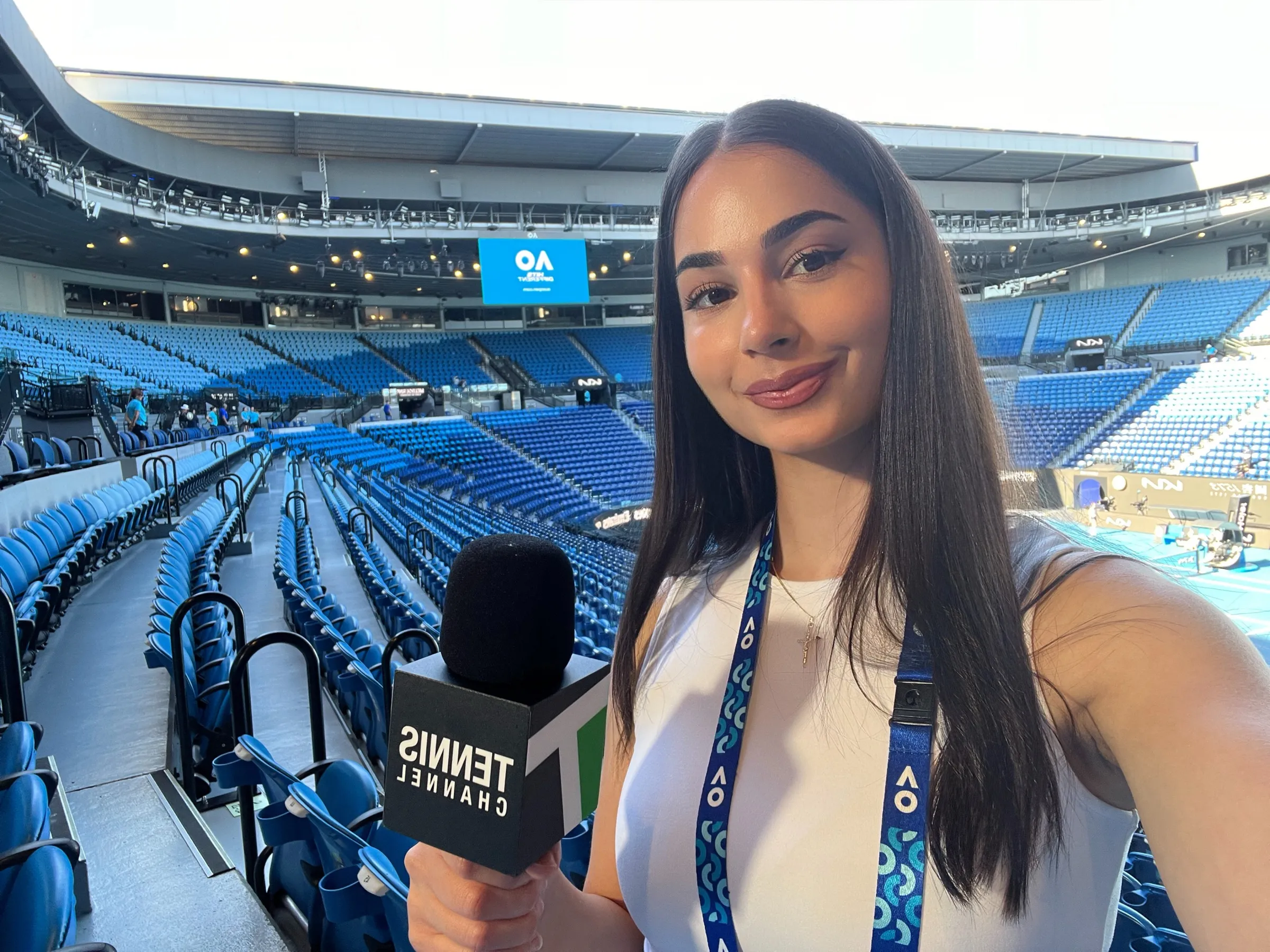 Maya smiling to camera in a large tennis stadium at the Australian Open, holding a Tennis Channel microphone.